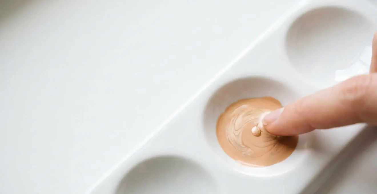 Vogelperspektive auf eine saubere weisse Oberfläche, auf der getönte Creme mit einem Tropfen Luminizer sanft gemischt wird